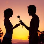 A romantic couple with the man giving the woman a flower with a colourful sunset behind them